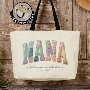 Pode incluir: Uma bolsa tote de lona na cor creme com al&ccedil;as pretas. A frente apresenta a palavra NANA em letras de tecido grandes e multicoloridas com v&aacute;rios padr&otilde;es. Abaixo, os nomes amelia, evan e sophia est&atilde;o impressos com est. 2026.