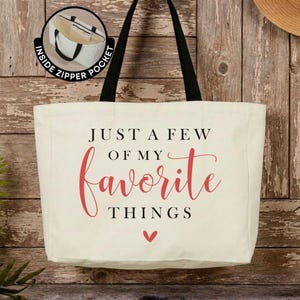 May include: A cream-colored canvas tote bag with black shoulder straps. The front features the black text "JUST A FEW OF MY" above the red script word "favorite" and the black text "THINGS" with a small red heart icon below the text.