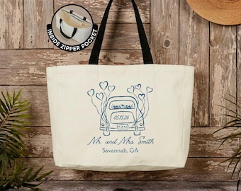 Può includere: Una borsa tote in tela color crema con spallacci neri. La parte anteriore presenta un disegno al tratto blu di un'auto con palloncini a forma di cuore, la data 05.15.26, il testo Mr. and Mrs. Smith e Savannah, GA. Un inserto mostra una tasca interna con cerniera.