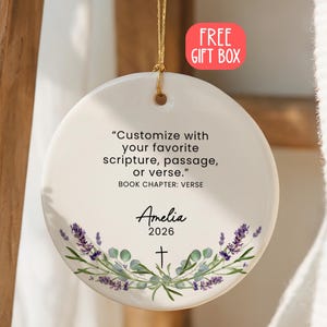 May include: A round white ceramic ornament hanging by a gold string. The text reads: Customize with your favorite scripture, passage, or verse. BOOK CHAPTER: VERSE. Below is the name Amelia, the year 2026, a small cross, and lavender sprigs.