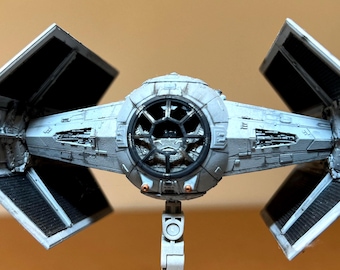 May include: A detailed model of a TIE fighter spacecraft, featuring a central spherical cockpit and two solar panel wings. The model is primarily gray with black accents, showcasing intricate mechanical details. The wings are angled, and the model is mounted on a white stand.