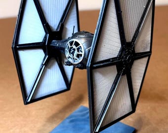 May include: A miniature TIE fighter model, featuring black solar panels and a central cockpit. The model is mounted on a blue base, showcasing the iconic design from the Star Wars franchise. The fighter's wings are angled, and the cockpit has intricate detailing.