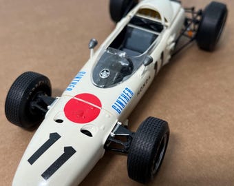 May include: A cream-colored vintage race car model with black tires and the number 11 on the front. The car features a red circle with a white center and the text "GINTHER" and "HONDA" in blue.