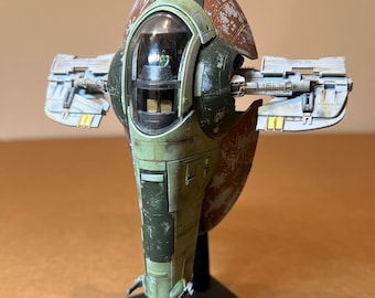 May include: A detailed model of a spaceship, featuring a weathered green, brown, and gray color scheme. The ship has a central cockpit with a clear dome, two side wings, and is mounted on a black stand. The model appears to be a replica from a science fiction franchise.