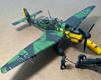 May include: A detailed model of a German WWII Stuka dive bomber, featuring green and black camouflage, yellow accents, and a black propeller. Miniature figures are shown working on a bomb. The model is a detailed replica of the aircraft.