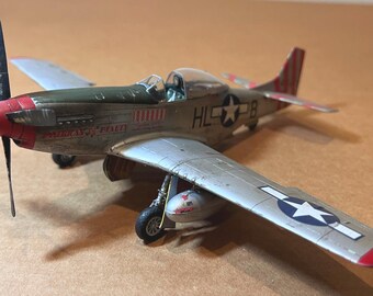 May include: A model airplane with a red nose and propeller, olive green and silver body, and red and white striped tail. The wings feature a white star in a blue circle. The plane has the text "AMERICAN BEAUTY" and "HL*28" on the body.