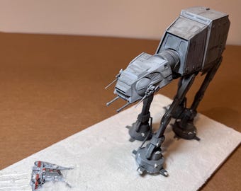 May include: A gray AT-AT model with detailed features, standing on a white surface. A smaller, red and white vehicle is positioned in front of the AT-AT, suggesting a battle scene. The model is a detailed replica of a Star Wars vehicle.