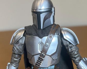 May include: A detailed action figure of a character from a science fiction series. The figure is clad in silver armor, including a helmet, chest plate, and shoulder pads. A black cape and strap are also visible. The figure is posed against a neutral background.
