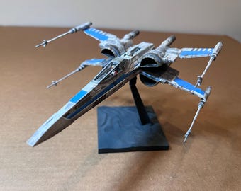 May include: A detailed model of a Star Wars X-wing fighter. The model is primarily white with blue accents on the wings and fuselage. It is mounted on a black base, showcasing the iconic design of the spacecraft.