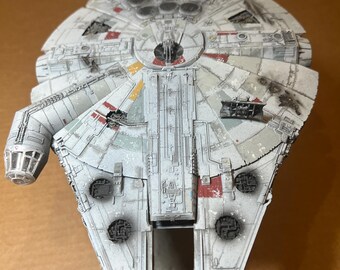 May include: A detailed model of the Millennium Falcon starship, featuring a weathered, gray and white color scheme with red, yellow, and black accents. The model has intricate surface details and is set against a neutral background.