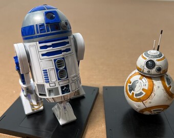 May include: Two Star Wars droid figures, R2-D2 and BB-8, are displayed on black bases. R2-D2 is white, blue, and silver, while BB-8 is white, orange, and silver. The droids are positioned against a neutral background.