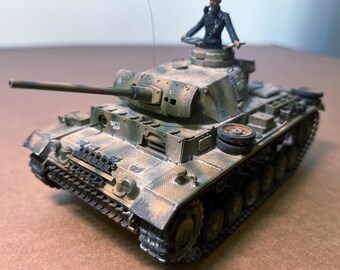 May include: A detailed model of a military tank in a camouflage pattern of brown, tan, and green. The tank has a long gun barrel and a small figure of a soldier in a black uniform. The tank's wheels and tracks are visible.