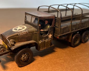 May include: A detailed model of a military truck, painted in olive drab. The truck features a canvas-covered bed, a driver figure, and the text "U.S.A. 4262921-S" with a star emblem on the hood. The tires are black.