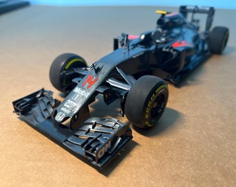 May include: A detailed model of a Formula 1 race car. The car is primarily dark gray with red and yellow accents. The number 14 is visible on the front. The tires have yellow lettering. The front wing is detached.