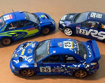 May include: Three blue and navy rally race cars with sponsor decals and racing numbers. The cars feature the Subaru and Ford Focus brands. One car has the number 1, another has 23, and the third has 25. The cars are on a brown surface.