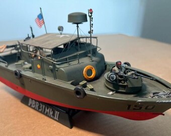 May include: A detailed model of a military patrol boat, painted in olive green with a red waterline. Features include a small American flag, a radar dome, and a machine gun turret. The model is labeled "PBR31MK.II" and has the number "150" on the bow.