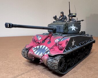May include: A detailed model of a military tank, painted in red and black with a shark mouth design on the front. The turret features the text "CE'S RED DEVILS" and a white star. Two figures are visible in the open turret.
