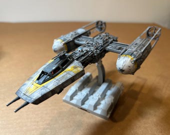 May include: A detailed model of a Y-wing starfighter, featuring a gray and yellow color scheme with weathering effects. The model is mounted on a gray base and has intricate detailing, including engine components and cockpit windows.