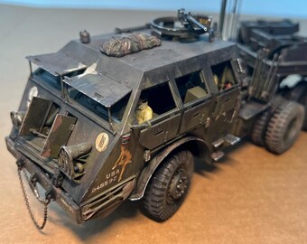 May include: A detailed model of a military truck, painted in shades of green and brown, with visible weathering. Features include a machine gun, open hatches, and the text "USA 545592". The model has multiple wheels and a chain.