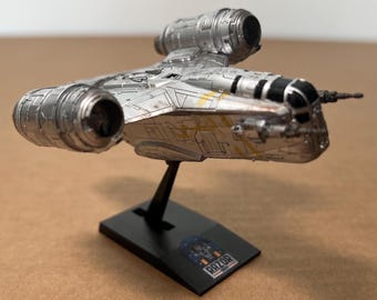 May include: A silver Razor Crest model spaceship with detailed paneling, yellow accents, and black detailing. The model is mounted on a black stand with the text "RAZOR CREST". The spaceship has two large engine components.