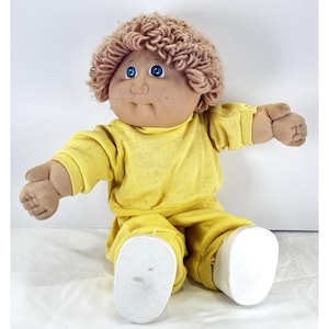 May include: A vintage Cabbage Patch Kid doll with a tan complexion, blue eyes, and light brown curly hair. The doll is wearing a yellow long-sleeved shirt and matching pants. The doll's feet are white.