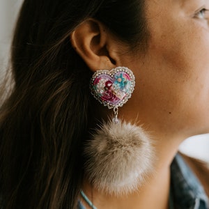 May include: A pair of heart-shaped earrings with a beaded design and a fluffy pom-pom dangle. The earrings are pink, purple, and blue with silver accents.