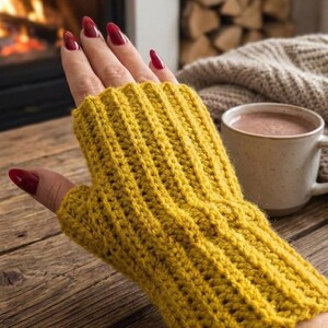 Könnte beinhalten: Ein senfgelber Fingerhandschuh aus Rippstrickstoff wird auf einer Holzoberfläche gezeigt. Der Handschuh wird an einer Hand mit rotem Nagellack getragen. Eine Tasse heiße Schokolade und ein Kamin befinden sich im Hintergrund.