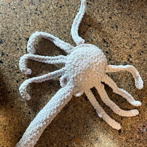 May include: A light gray crocheted octopus toy with eight long, curved tentacles. The octopus has a round head and the yarn has flecks of multiple colors. The toy is made of a soft, plush material.