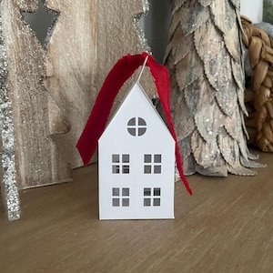 Può includere: Ciondolo a forma di casa in carta bianca con nastro rosso. La casa ha finestre ritagliate e un cerchio al centro.