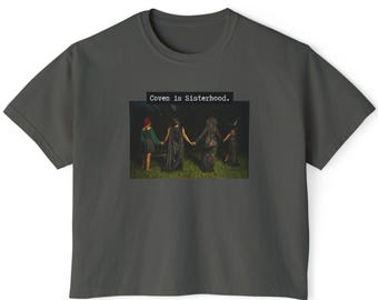 May include: Dark gray t-shirt featuring a rectangular graphic with four figures holding hands. The figures are dressed in dark clothing and standing on grass. The text "Coven is Sisterhood." is above the graphic.