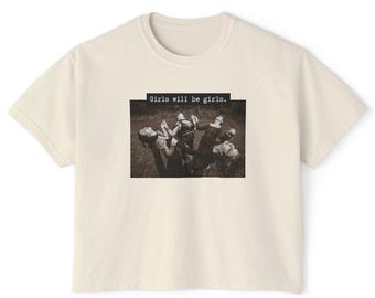 May include: Cream-colored t-shirt with a crew neck. The shirt features a sepia-toned rectangular graphic of people in a field, with the text "Girls will be girls." above the graphic. The shirt is made of a soft material.