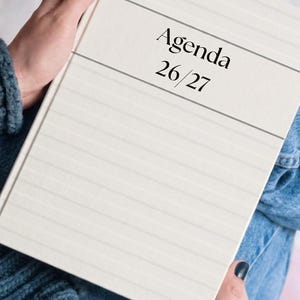 Puede incluir: Una agenda de tapa dura color crema con líneas horizontales en relieve en la cubierta. El texto "Agenda 26/27" está impreso en negro en el centro. El planificador tiene un diseño limpio y estructurado con un tono neutro y acabado mate.
