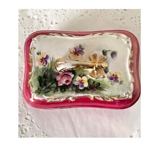May include: A rectangular trinket box with a pink rim and a floral design. The lid features a gold-colored handle and a painted scene of pink roses, purple pansies, and green foliage on a white background. The box has gold accents.