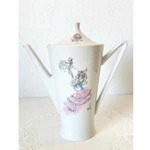 May include: White porcelain coffee pot with a tall, slender design and a decorative handle. The pot features a lid and is adorned with a colorful illustration of a woman in a pink dress. The design is reminiscent of vintage or antique tableware.