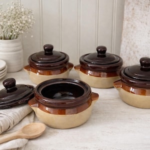May include: A collection of four ceramic, two-tone brown and beige lidded pots, with a fifth pot open, revealing the interior. A wooden spoon and a stack of white plates are also visible.