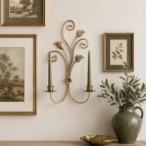 May include: A gold-toned metal wall sconce featuring two candle holders and a decorative vine-like design with leaf accents. The sconce holds two slender, olive-green taper candles, creating a symmetrical arrangement on the wall surface.