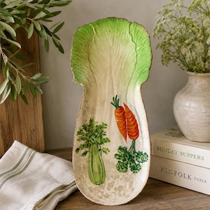 May include: A ceramic serving platter shaped like a large lettuce leaf with a cream-colored base. The platter features painted illustrations of two orange carrots and two green celery stalks on the surface, with a vibrant green leaf edge.