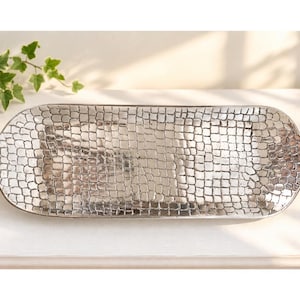 May include: A long, rectangular silver-toned metal tray with rounded ends and integrated handles. The surface features a raised, repeating grid pattern that resembles cobblestones or scales, creating a textured, reflective metallic finish.