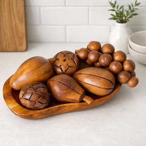 May include: A wooden decorative tray filled with carved wooden fruit. The tray holds a variety of fruits, including pears, grapes, and other stylized shapes. The fruit is a warm brown color, and the tray has a natural wood grain.