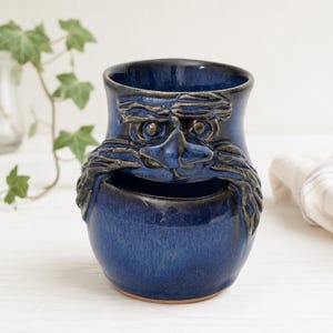 May include: A dark blue ceramic mug featuring a sculpted face with prominent eyebrows, a nose, and a mustache. The glaze has a glossy finish with darker blue and black tones. The mug has a wide opening at the top and a secondary opening below the mustache.