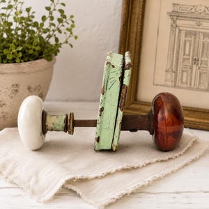 May include: A door handle set featuring a white porcelain knob on one side and a dark brown glass knob on the other, connected by a metal spindle through a rectangular, distressed mint green metal backplate with visible rust and paint wear.