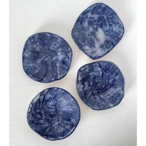 May include: Four small, irregularly shaped ceramic bowls featuring a glossy finish with a marbled pattern of deep navy blue and white. Each bowl has a unique, organic silhouette with slightly raised, uneven edges and a smooth, reflective surface.