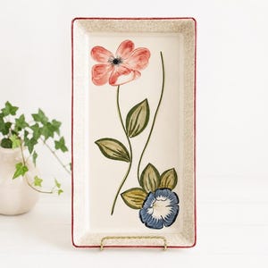 May include: A rectangular ceramic serving platter with a cream-colored center featuring a hand-painted coral pink flower and a smaller blue flower with green leaves. The platter has a speckled beige border and a thin red rim around the edge.
