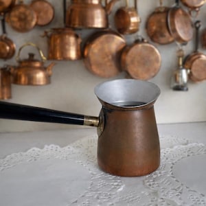 May include: A copper coffee pot with a black handle sits on a white lace doily. The pot has a silver rim and is surrounded by other copper kitchenware hanging in the background. A classic kitchenware item.