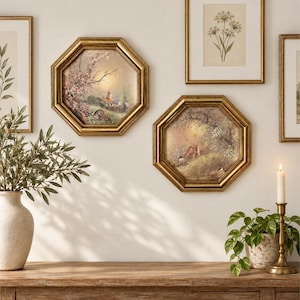 May include: Two octagonal wall art pieces in gold frames featuring landscape paintings of rural scenes with trees and buildings. The artwork is displayed alongside rectangular framed prints, a ceramic vase, a plant, and a candle holder.