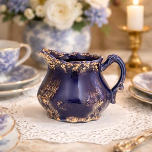 May include: A navy blue ceramic creamer with a gold-speckled rim and accents. The creamer has a curved handle and a scalloped edge. It is part of a tea set, with other pieces visible in the background.