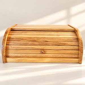 May include: A rectangular wooden bread box featuring a roll-top lid with horizontal slats and a small, centered round knob. The container is crafted from light-toned wood with a visible natural grain pattern, showcasing a smooth, polished finish.