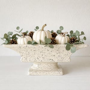 May include: A cream-colored ceramic pedestal bowl with a black speckled finish, filled with three small white pumpkins, several brown pinecones, and green eucalyptus sprigs arranged in a decorative display for a tabletop centerpiece.