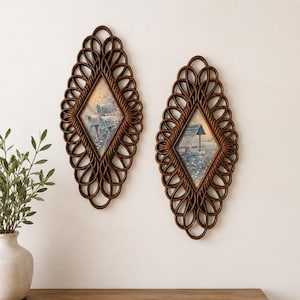 May include: Two oval-shaped wall hangings featuring dark brown, woven rattan frames with intricate, looping patterns surrounding a central diamond-shaped panel. Each panel contains a printed illustration of a rural scene with a small structure.