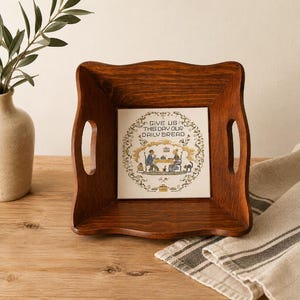 May include: A square wooden serving tray with cutout handles and a dark brown finish. The center features a white tile with a decorative border and the text "GIVE US THIS DAY OUR DAILY BREAD" above an illustration of two people at a table.
